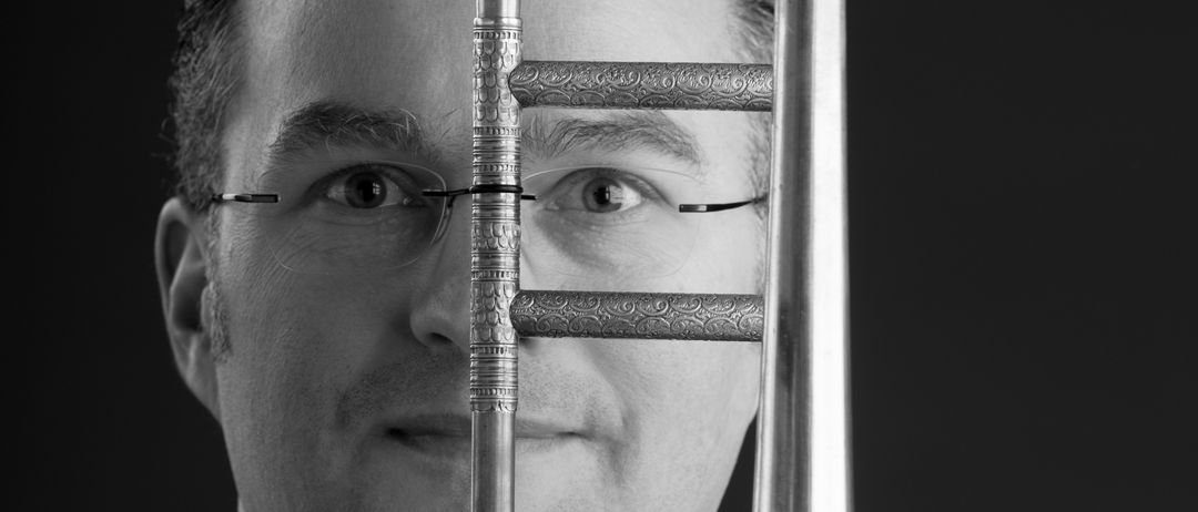 Ein schwarz-weißes Foto eines Mannes mit Brille, der ein Posaune hält. Die Posaune ist über dem Gesicht des Mannes positioniert.