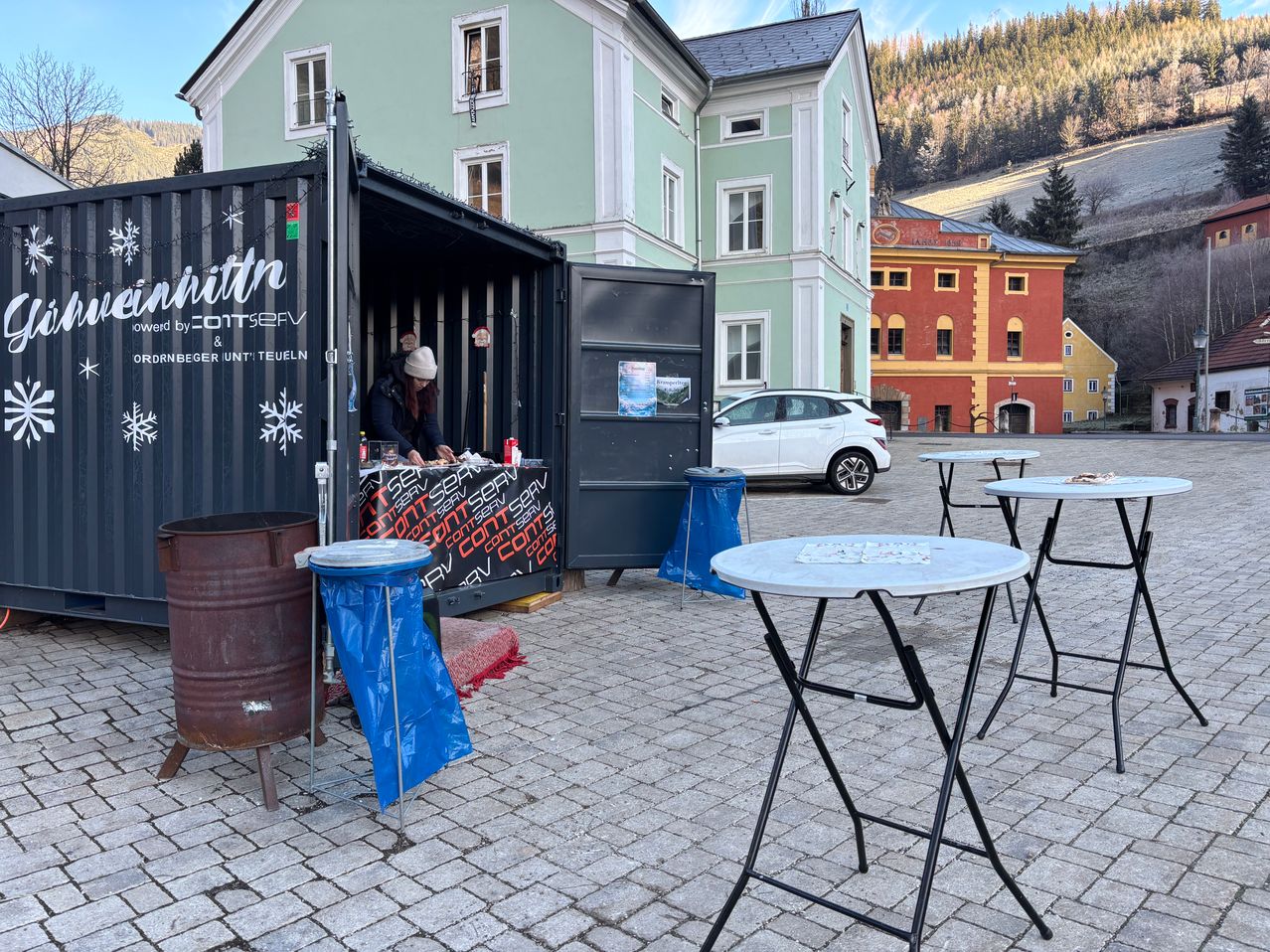 Ein Container-Shop ist vor einem Gebäude mit rot-gelber Fassade geöffnet. Eine Person ist drinnen und draußen gibt es Tische und einen Mülleimer. Ein weißes Auto ist in der Nähe geparkt.