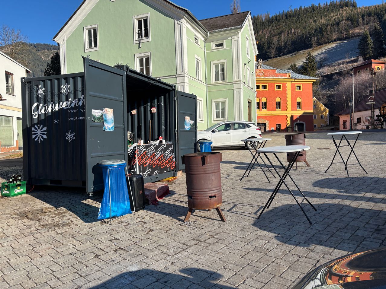 Ein Container-LKW steht vor einem Gebäude mit einem roten Fass und Tischen. Dahinter befinden sich zwei Gebäude, eines grün und eines orange. Ein weißes Auto ist vor dem orangen Gebäude geparkt.