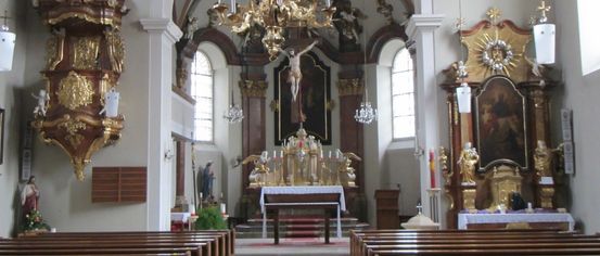 The interior of a church, featuring a large gold chandelier above the altar, statues, and intricate decorations. Pews line both sides of the nave.