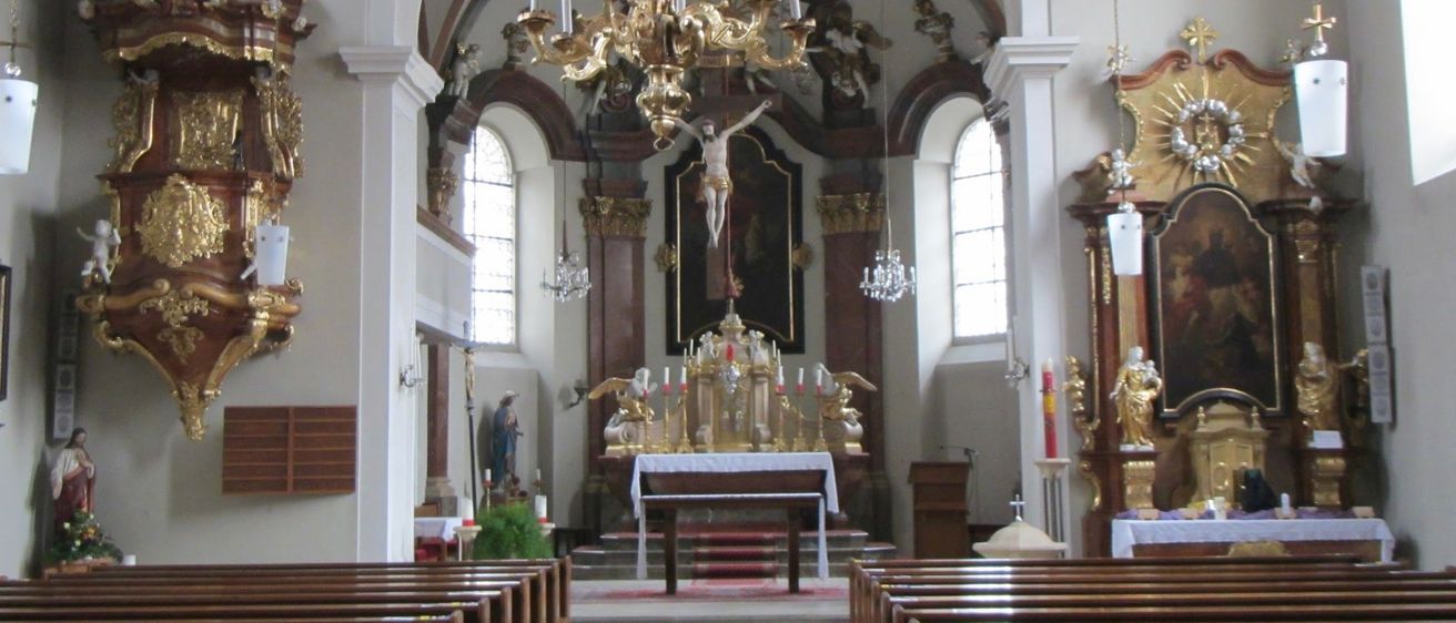 Das Innere einer Kirche mit einem großen goldenen Kronleuchter über dem Altar, Statuen und aufwendigen Verzierungen. Bänke säumen beide Seiten des Kirchenschiffs.