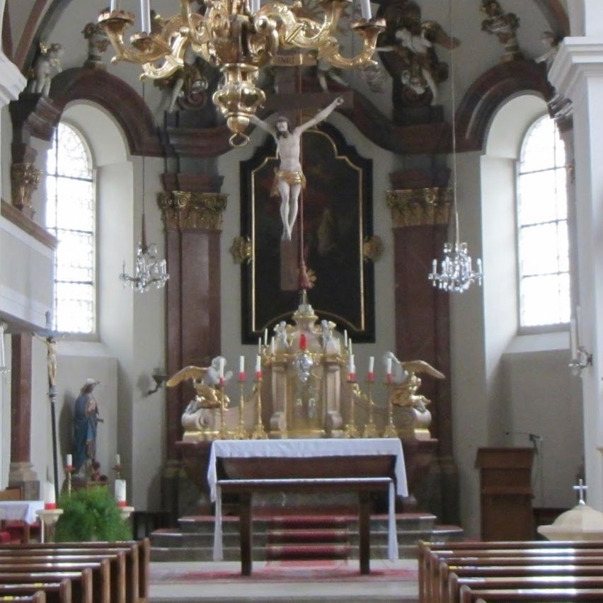 Das Innere einer Kirche mit einem großen goldenen Kronleuchter über dem Altar, Statuen und aufwendigen Verzierungen. Bänke säumen beide Seiten des Kirchenschiffs.