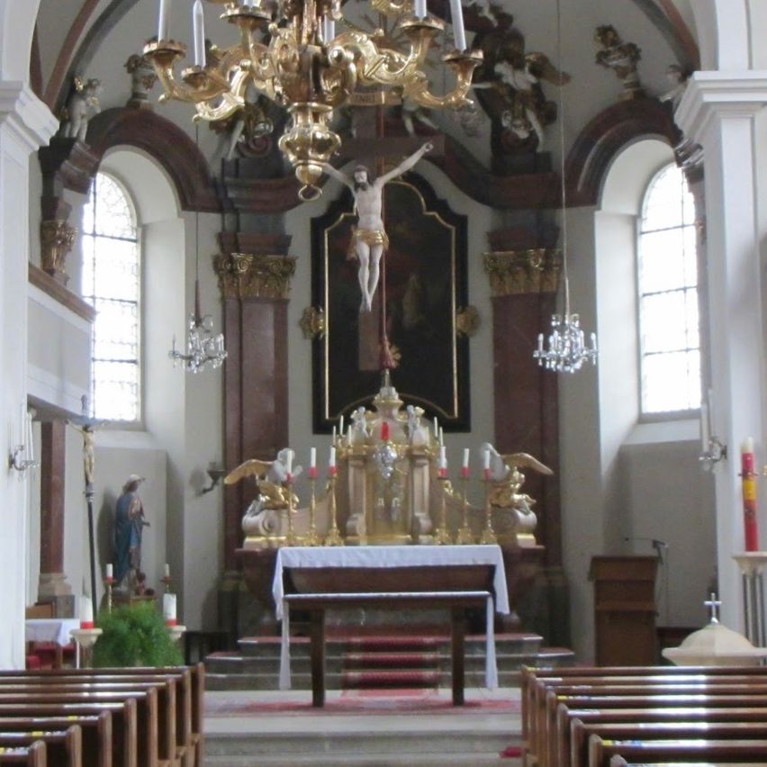 The interior of a church, featuring a large gold chandelier above the altar, statues, and intricate decorations. Pews line both sides of the nave.