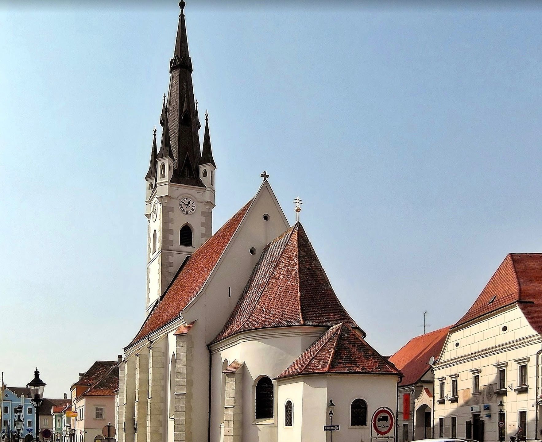 Eine gotische Kirche mit einem hohen Turm und roten Dächern, vor einem klaren blauen Himmel. Eine Uhr ist am Turm sichtbar.