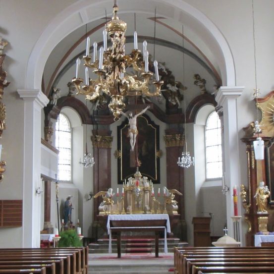 Der Innenraum einer Kirche mit einem aufwendigen Altar, einem großen goldenen Kronleuchter, einem Kruzifix und einer Statue von Jesus am Kreuz.