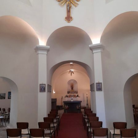 Eine leere Kirche mit gewölbten Wänden und einem Kreuz über dem Altar. Ein roter Teppich führt zum Altar, und Stühle sind auf beiden Seiten angeordnet. Gerahmte Bilder und Kerzen schmücken die Wände.