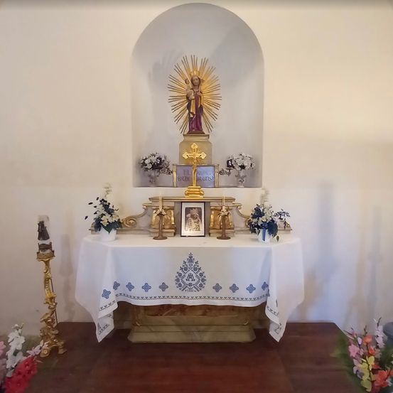 Ein religiöser Altar mit einer Statue der Jungfrau Maria. Ein weißer Stoff mit blauen Mustern liegt auf dem Altar. Ein goldenes Kreuz, Blumen und ein Bilderrahmen stehen auf dem Altar. Zwei goldene Kerzen stehen auf beiden Seiten des Rahmens.
