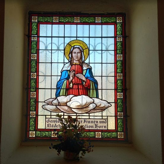 Gerahmtes Buntglasfenster mit Maria, der Mutter Jesu, die eine Wolke hält, mit Blumen darunter.