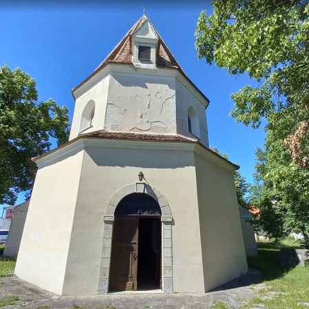 Eine alte weiße Kapelle mit einem braunen Dach, einem Rundbogeneingang und einer Lampe. Sie ist von grünen Bäumen und Gras umgeben an einem sonnigen Tag.