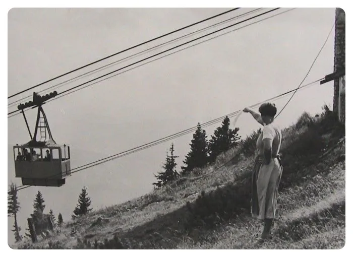 Eine Frau steht auf einem Hang und hält ein Seil einer Seilbahn, während eine Seilbahn hinter ihr aufsteigt, umgeben von Bäumen und Gräsern.