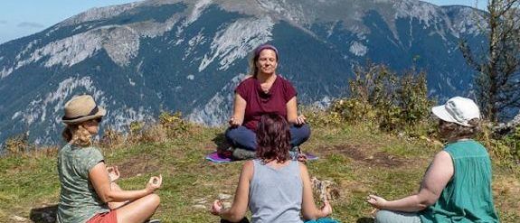 Vier Frauen sitzen in einer Yoga-Pose auf einem grasbewachsenen Berghang mit einer Kulisse von Bergen und blauem Himmel.