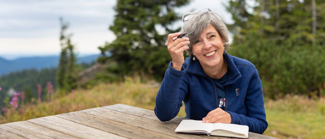 Eine ältere Frau mit grauem Haar sitzt auf einer Holzbank und lächelt. Sie hält einen Stift und ein offenes Buch, mit Kiefern im Hintergrund.