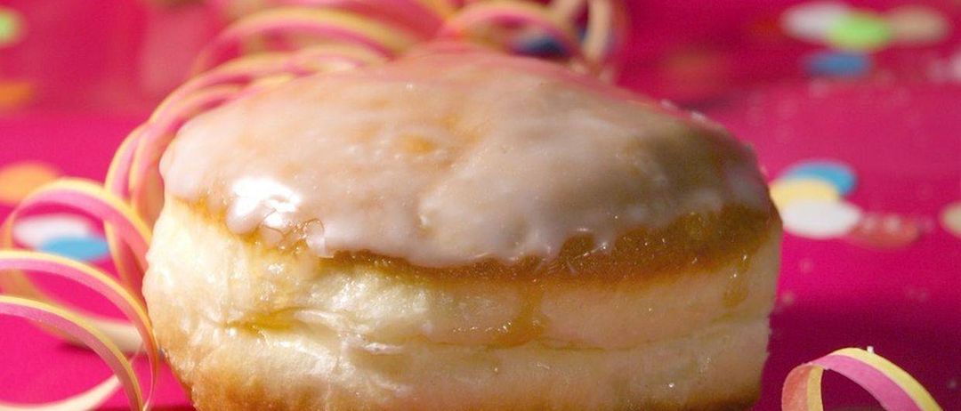 Ein Nahaufnahme eines glasierten Donuts mit Streuseln liegt auf einer rosa Oberfläche, umgeben von bunten Luftschlangen und Konfetti.