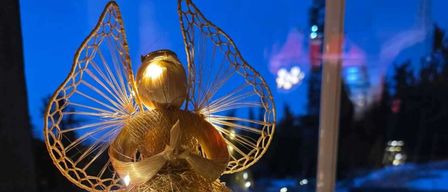 Eine goldene Engel-Dekoration mit Flügeln und Lichtern steht auf einer Fensterbank mit Nachtblick. Ein Kranz mit Lichtern ist in der Nähe.