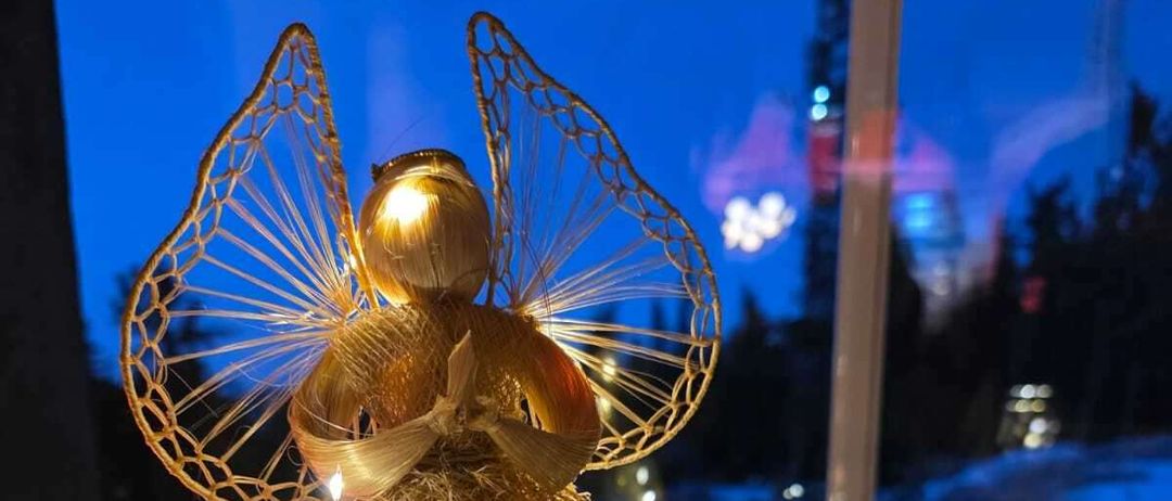 Eine goldene Engelsfigur mit beleuchteten Flügeln steht auf einem Tisch, erleuchtet von Lichtern. Daneben befindet sich ein Weihnachtskranz. Der Blick aus dem Fenster zeigt eine Winterlandschaft bei Nacht.