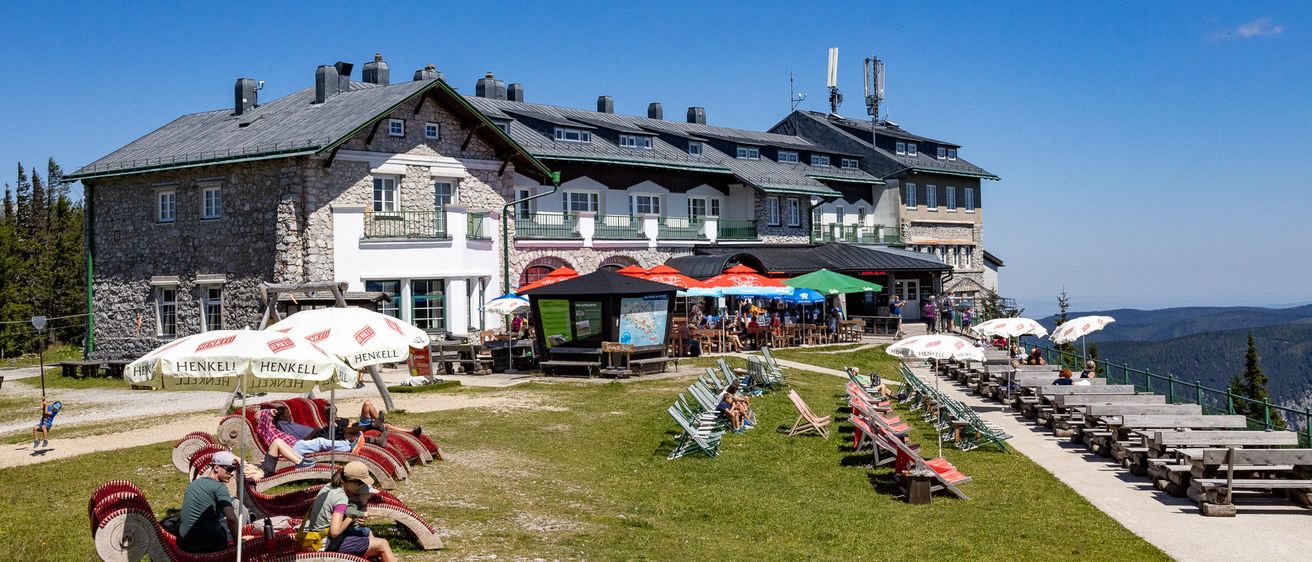 Ein sonniger Tag in einem Bergresort, wo Menschen die Sitzgelegenheiten im Freien, ein großes Gebäude und bunte Sonnenschirme genießen.