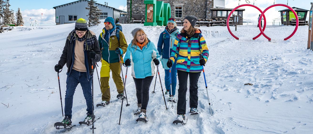 Sechs Personen gehen auf Schneeschuhen vor einem Steingebäude mit der Nummer 40. Dahinter steht ein großes Gebäude mit vielen Fenstern.