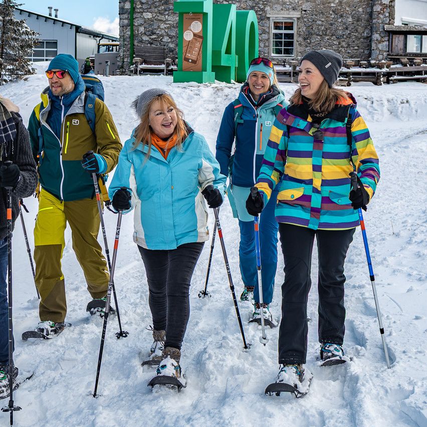 Sechs Personen gehen auf Schneeschuhen vor einem Steingebäude mit der Nummer 40. Dahinter steht ein großes Gebäude mit vielen Fenstern.