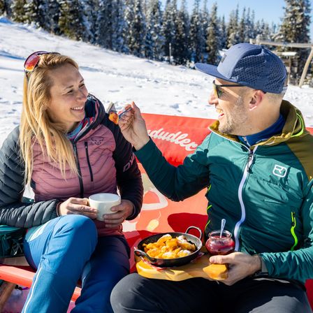 Ein Paar genießt eine Mahlzeit auf einer Bank in einer verschneiten Landschaft, wobei der Mann der Frau Essen reicht.