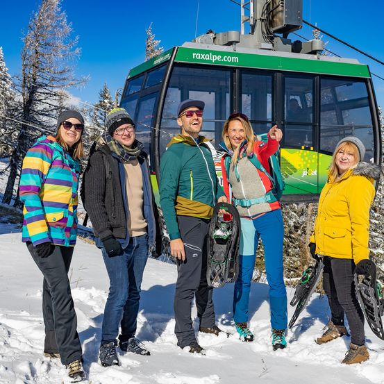 Fünf Personen, gekleidet in Winterkleidung, stehen im Schnee vor einer Gondel. Die Gondel hat einen grünen Rahmen mit 'raxalpe.com' darauf. Sie alle lächeln und scheinen für ein Foto zu posieren.