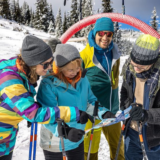 Eine Gruppe von vier Personen in Winterkleidung steht im Schnee und betrachtet eine Karte. Sie lächeln und scheinen ihre Route zu planen. Hinter ihnen ist ein großer roter Bogen und verschneite Bäume zu sehen.