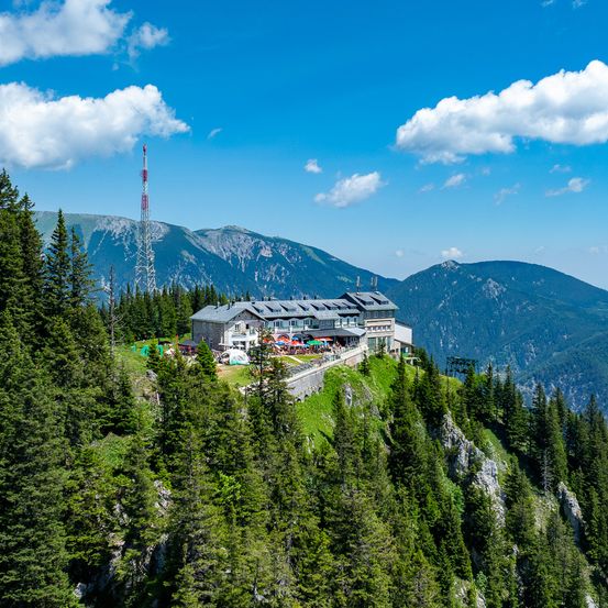 Ein Gebäude auf einem Hügel mit einem Turm, umgeben von Bäumen und Bergen unter einem blauen Himmel.