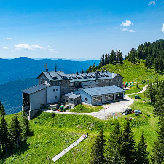 Eine Berghütte mit Solarpanelen auf dem Dach, umgeben von grünem Gras, Bäumen und einer Bergkette.