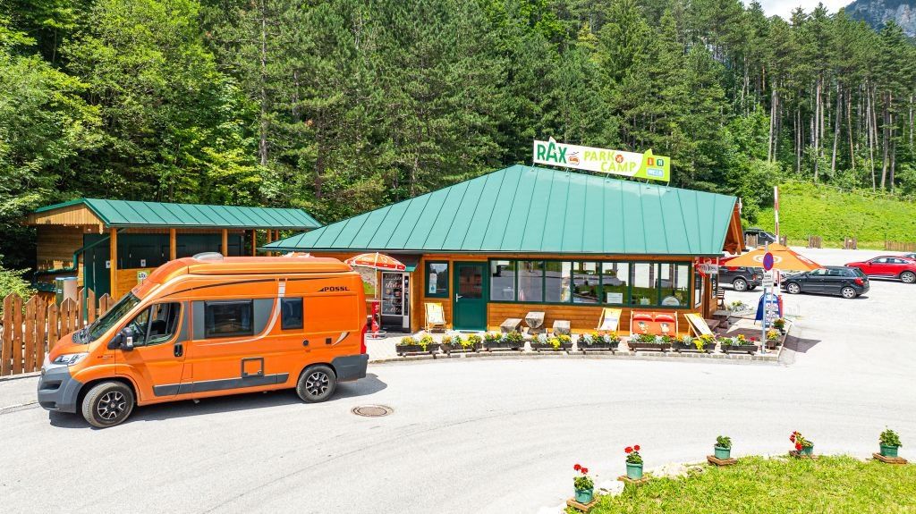 Ein orangefarbener Wohnmobil steht vor dem RAX Park & Camp, das ein grünes Dach hat und von üppiger Grünanlage umgeben ist.