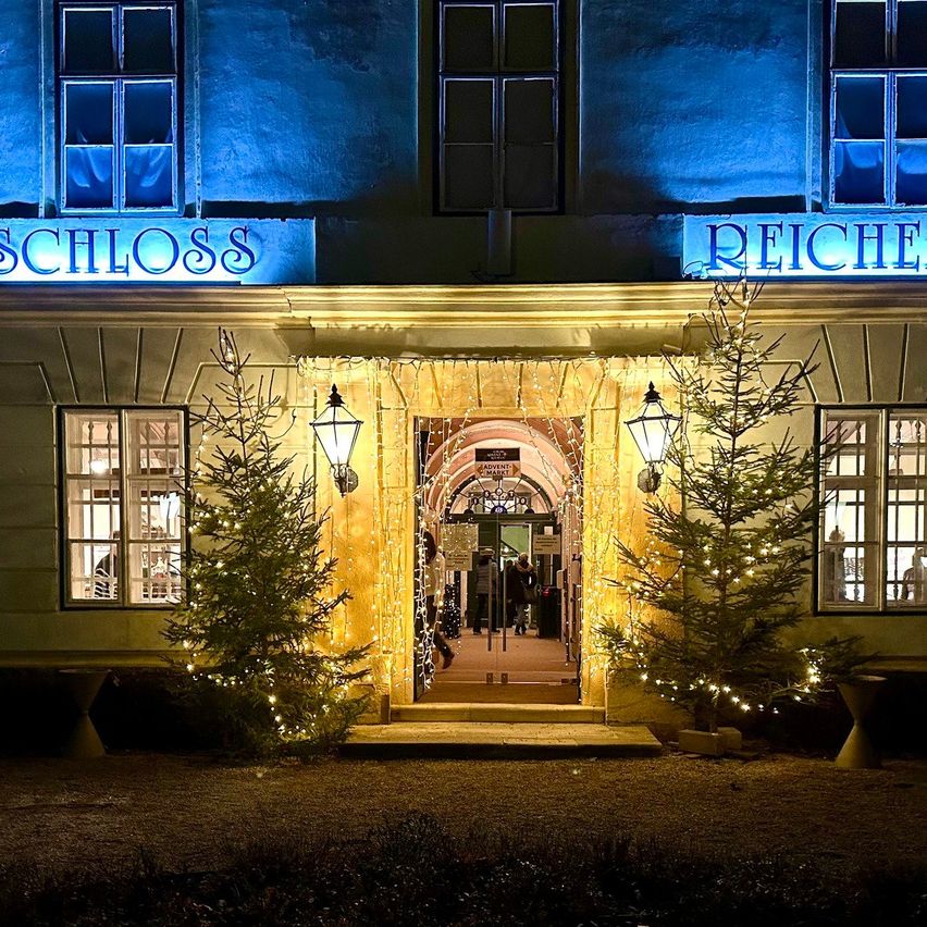 Der Haupteingang des Reiche Schloss ist mit Weihnachtsbäumen und Lichtern geschmückt. Das Gebäude hat blaue Beleuchtung oben und an den Fenstern. Es gibt eine Lampe auf beiden Seiten des Eingangs.