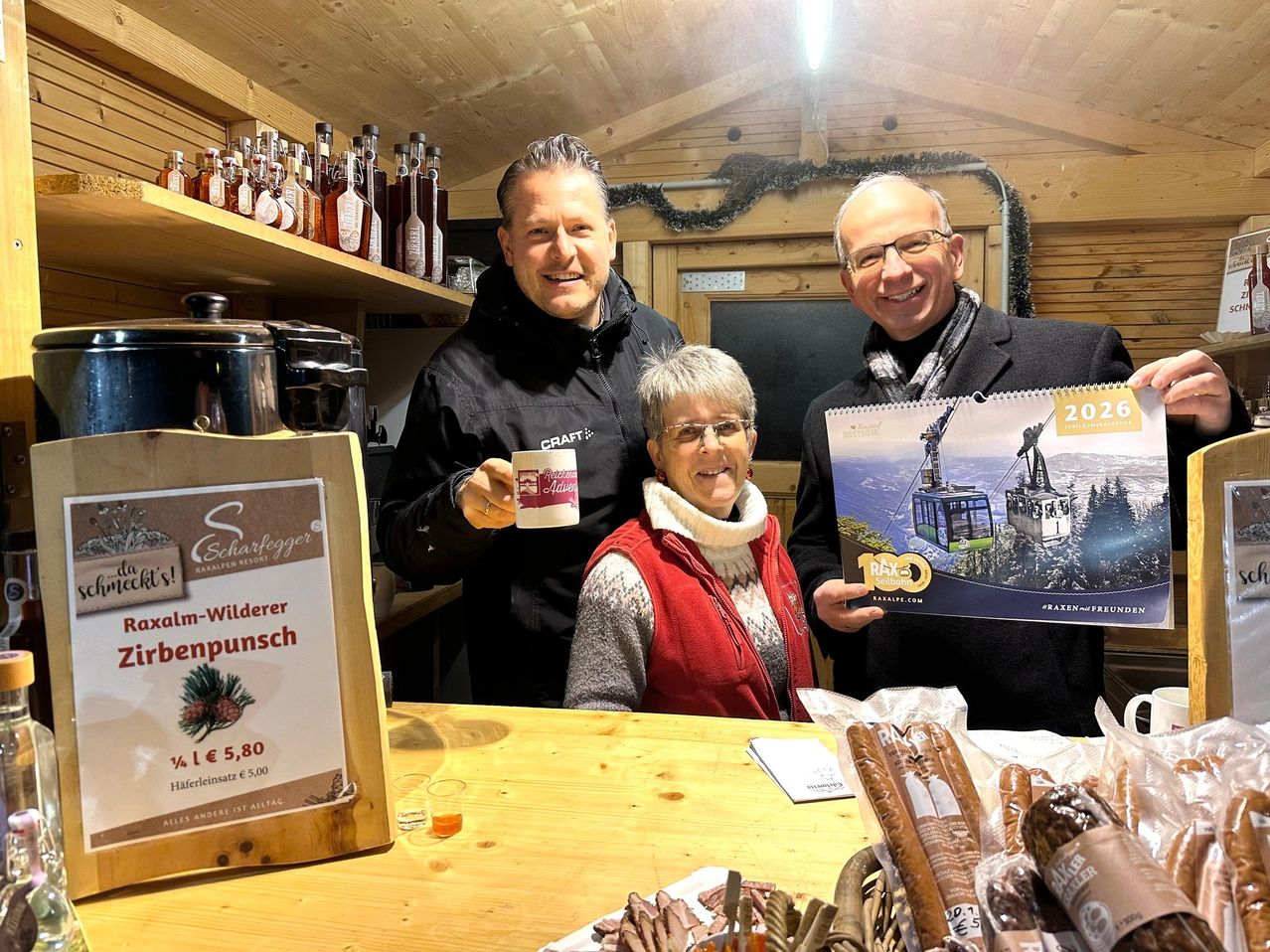 Drei Menschen posieren für ein Foto in einer Hütte mit Holzregalen, einem Kalender und verschiedenen Gegenständen. Der Mann links hält eine Tasse, die Frau in der Mitte lächelt und der Mann rechts hält einen Kalender.