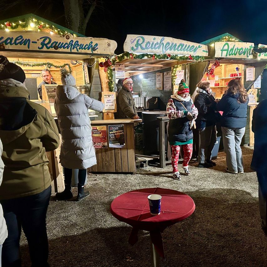 Menschen versammeln sich bei Nacht um Essensbuden, geschmückt mit Weihnachtslichtern. Eine Bude bietet Rauchkuche an, eine andere Reichenauver und eine dritte ist Advent beschriftet. Ein roter Tisch steht im Vordergrund.