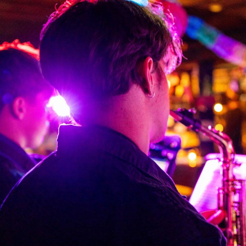 A man plays the saxophone under a spotlight, with another musician behind him in a dimly lit room with colorful lights.
