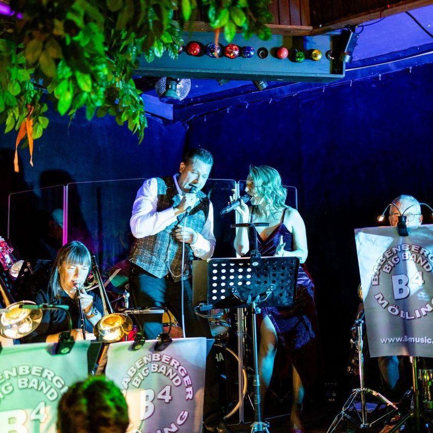 A band performs on stage with a blue backdrop, featuring a man and woman singing, a trombone player, and a drum set. A sign with 'B4' and a website is visible.