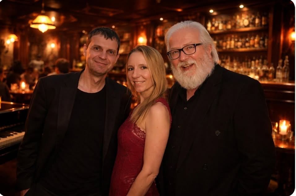 Three people are standing together in a bar, smiling for a photo. The man on the left is wearing a black suit, the woman in the middle is wearing a red dress, and the man on the right has a white beard and is wearing glasses.