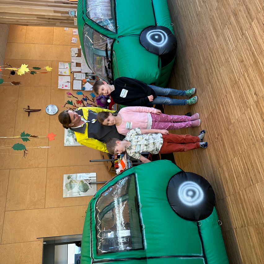 A group of children and an adult pose in front of inflatable green cars. The room has wooden floors and walls with decorations.