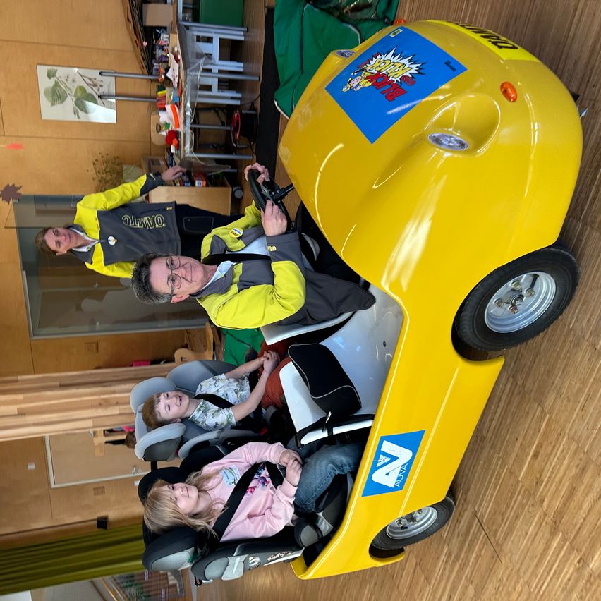 Three individuals, including two children, are enjoying a ride in a yellow, cartoon-themed vehicle. Two adults are standing behind the vehicle, one wearing a reflective vest. The scene takes place indoors, likely in a community center.