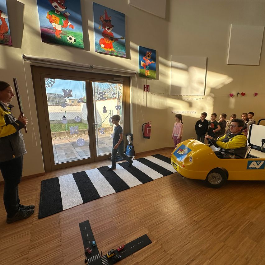 A man drives a yellow toy car on a track, while a woman stands nearby with a stick. A group of children stands on the side, watching. The room has a glass door, a fire extinguisher, and walls decorated with paintings.