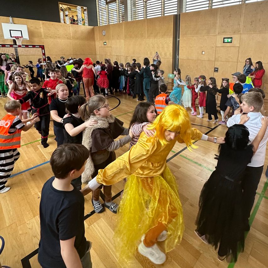 Eine große Gruppe von Kindern tanzt in einer Turnhalle, einige in Kostümen. Eine Person in einem gelben Kostüm tanzt mit anderen.