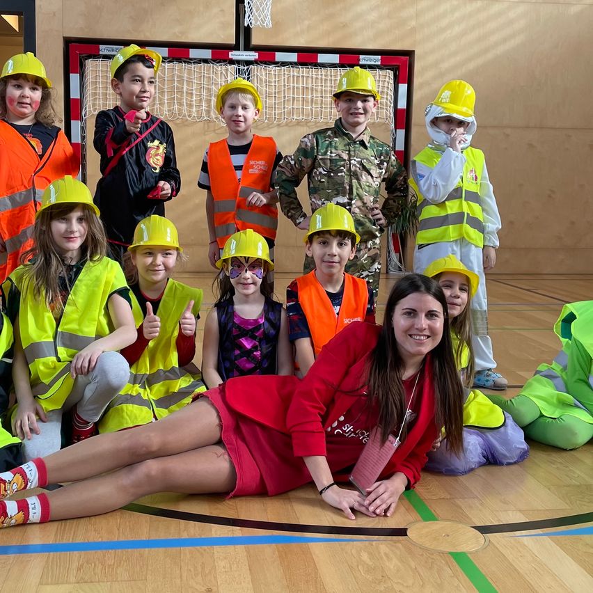 Gruppe von Kindern in verschiedenen Kostümen, darunter Bauhelme und Sicherheitswesten, posiert mit einer erwachsenen Frau in einer Turnhalle. Ein Basketballkorb ist im Hintergrund zu sehen.