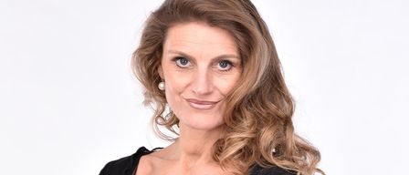 A woman with blonde wavy hair is wearing a black blouse with mesh sleeves. She is standing with her arms crossed and smiling. She has a gold watch and a ring on her left hand. She seems to be posing for a photograph.
