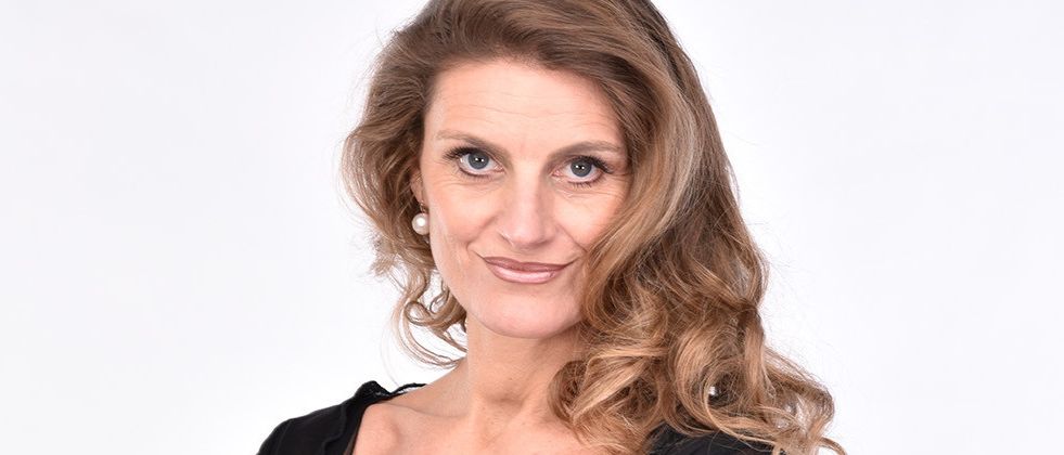 Eine Frau mit blonden welligen Haaren trägt ein schwarzes Oberteil mit Netzärmeln. Sie steht mit verschränkten Armen und lächelt. Sie hat eine goldene Uhr und einen Ring an ihrer linken Hand. Sie scheint für ein Foto zu posieren.