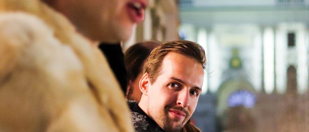 Zwei Männer, einer lächelnd, stehen vor einem Gebäude mit einer Kuppel, beleuchtet von Lichtern in der Nacht. Einer hält ein Papier.