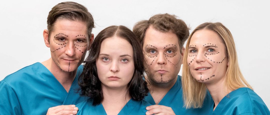 Eine Gruppe von vier Personen, gekleidet in blaue Kittel mit Gesichtsmarkierungen und -ausdrücken, posiert möglicherweise für ein Foto in einem medizinischen Umfeld.