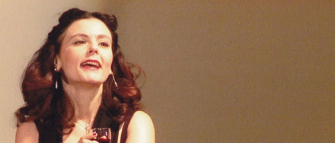 Eine Frau mit roten Haaren, die ein schwarzes Kleid und Handschuhe trägt, hält ein Weinglas und singt. Sie steht vor einem verschwommenen Publikum.