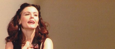 Eine Frau mit roten Haaren, die ein schwarzes Kleid und Handschuhe trägt, hält ein Weinglas und singt. Sie steht vor einem verschwommenen Publikum.