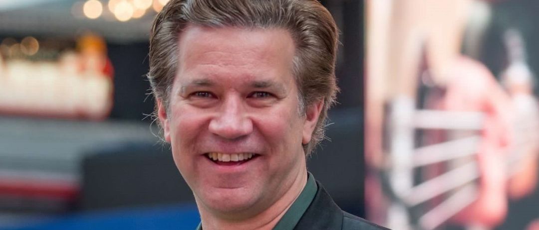 Ein Mann in einem schwarzen Anzug und grünem Hemd, lächelt und steht vor einem verschwommenen Hintergrund.