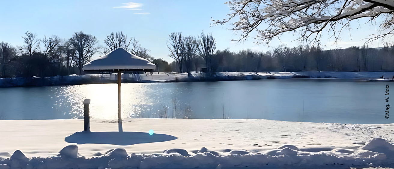 Eine Winterszene mit einem schneebedeckten Strandschirm am See, der Sonnenlicht auf dem Schnee reflektiert.