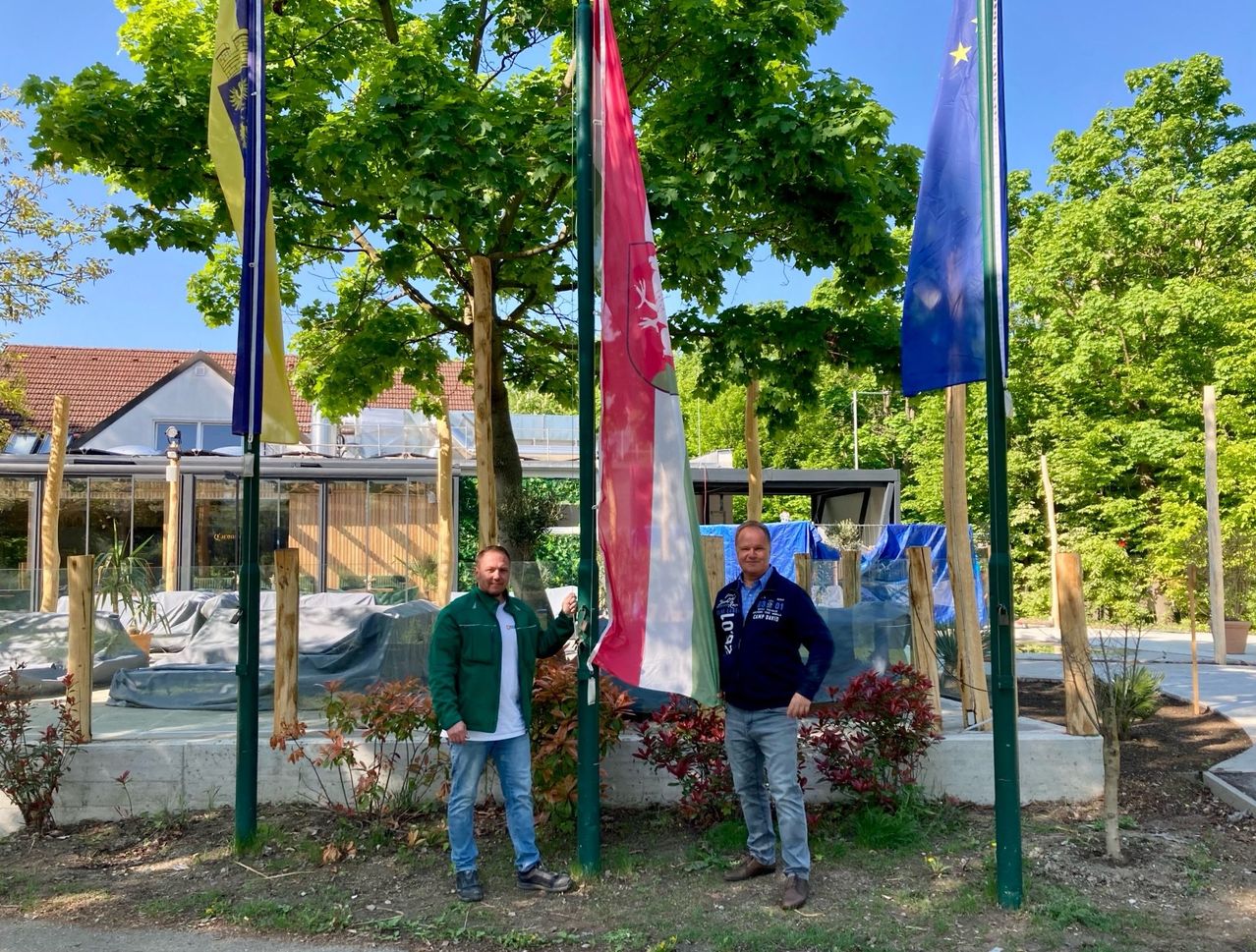 Zwei Männer stehen vor einem Fahnenmast, einer hält eine Flagge Deutschlands und der andere eine Flagge Italiens. Sie tragen beide Freizeitkleidung.
