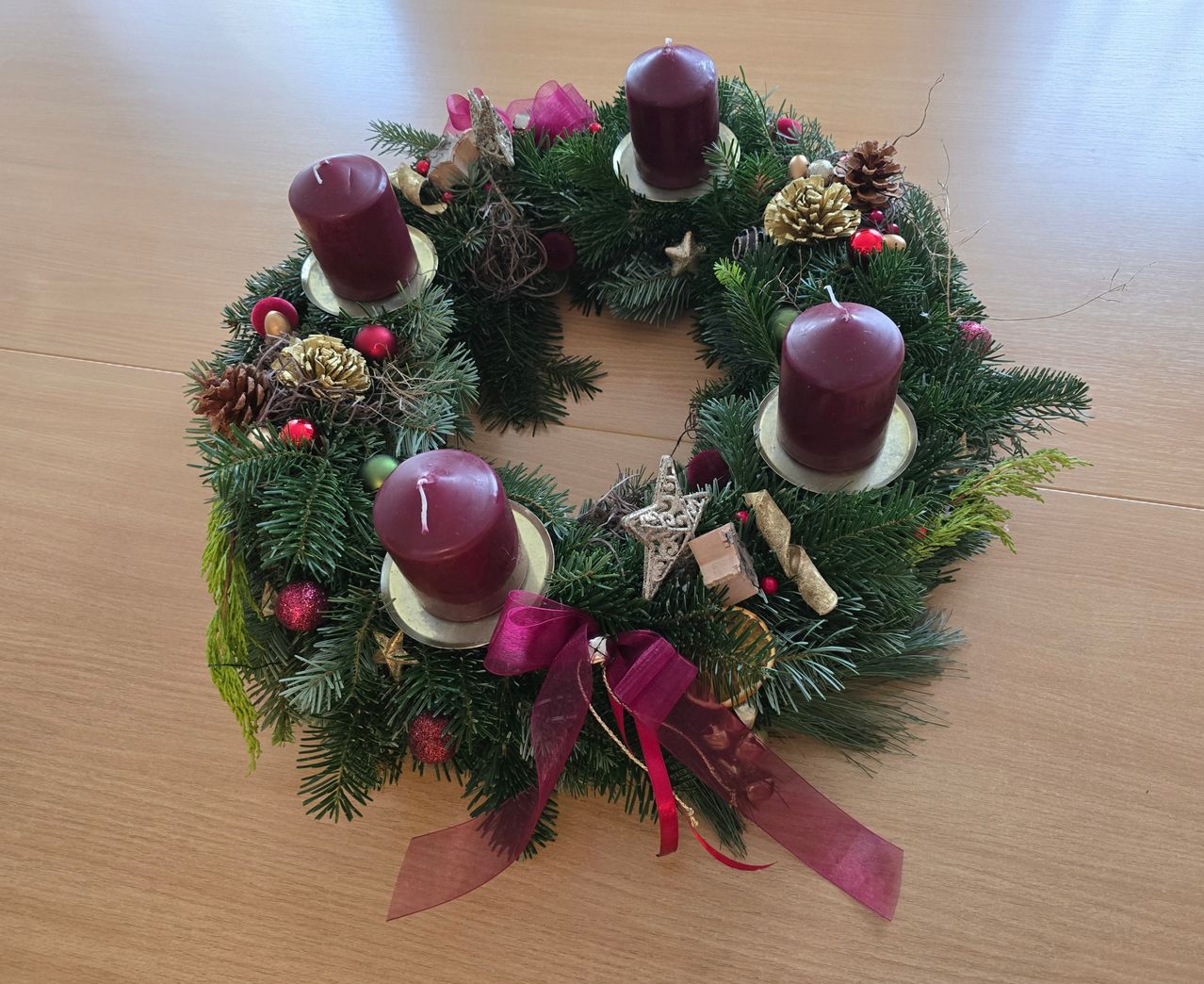 Ein Kranz mit vier roten Kerzen, geschmückt mit Tannenzapfen, roten Bändern und Ornamenten, ruht auf einer hölzernen Oberfläche.