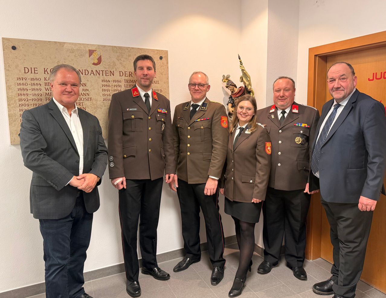 Sechs Personen, vier in Militäruniformen und zwei in Anzügen, stehen gemeinsam in einem Raum. Hinter ihnen befindet sich eine Plakette mit Namen und Jahren.